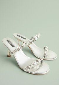 Silversandaler med klara remmar som har blommiga dekorationer. De har en elegant design och en blank finish, lämpliga för formella tillfällen.