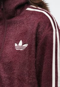 Close-up de uma pessoa a usar uma jaqueta peluda na cor bordô com o logo branco da Adidas e duas listras brancas nos ombros e nas mangas.