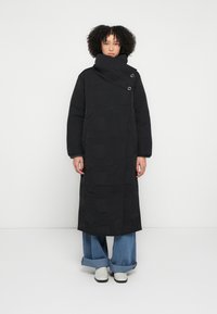 Black quilted long coat with geometric pattern, high collar, and two button accents; paired with wide-leg denim pants and white shoes.