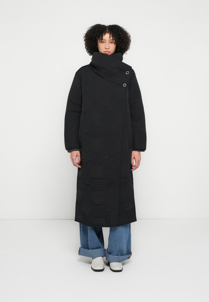 Black quilted long coat with geometric pattern, high collar, and two button accents; paired with wide-leg denim pants and white shoes.