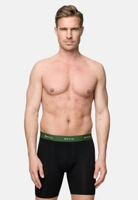 Schwarze Boxershorts mit grünem Bund, der weißen Logobeschriftung. Der Stoff wirkt glatt und eng anliegend. Das Modell zeigt eine neutrale Pose.