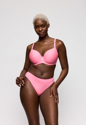 Vrouw met kort blond haar, gekleed in een bijpassende roze beha en hoge taille slip, staand tegen een effen grijze achtergrond.