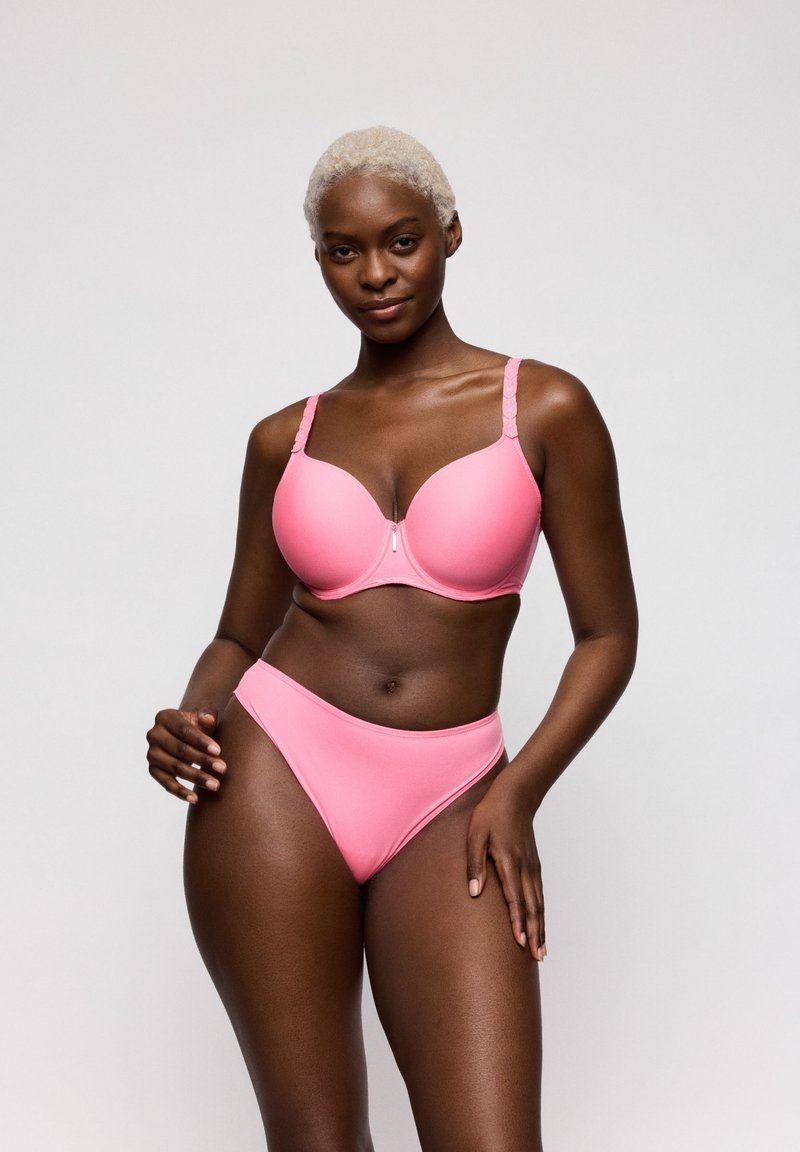 Donna con capelli corti biondi che indossa un reggiseno rosa coordinato e mutandine a vita alta, in piedi davanti a uno sfondo grigio uniforme.