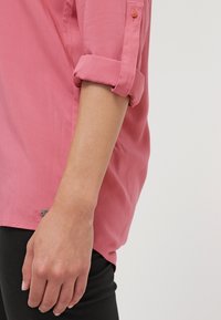 Bras d'une personne portant une chemise rose à manches longues retroussées et un pantalon noir, posé devant un fond clair uni.