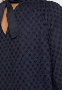 Blusa ricamata blu navy con piccoli motivi floreali, tessuto testurizzato e dettaglio con fiocco nella parte posteriore del colletto.