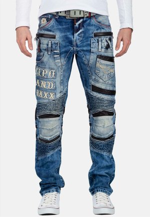 Mann trägt blaue, abgenutzte Jeans mit mehreren Taschen, Patches und dem Schriftzug „CIPO AND BAXX“, kombiniert mit weißen Sneakers und einem weißen Hemd.