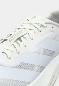 Weißer Sportschuh mit hellblauen Akzenten, ausgestattet mit einem strukturierten Mesh-Obermaterial, flachen Schnürsenkeln und einem auffälligen Markenetikett auf der Zunge.