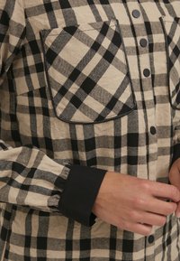 Brun och svart rutigt flanellskjorta med en bröstficka och svarta manschetter; har knappknäppning fram och mjuk textur.