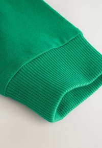 Manga de algodón verde con puño acanalado, textura suave y borde redondeado. La tela tiene un tono vibrante y un patrón de tejido uniforme.