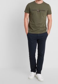 Mandlig model iført olivengrøn Tommy Hilfiger t-shirt, mørkeblå bukser og hvide sneakers, stående med hænderne i lommerne på grå baggrund.