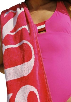 Costume da bagno rosa con dettaglio cut-out, abbinato a un asciugamano rosa e bianco a fantasia drappeggiato sulla spalla. Texture morbida visibile.