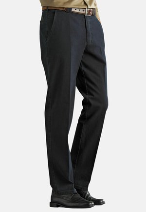 Mann trägt schwarze gerade geschnittene Hose, braunen Ledergürtel, beiges Hemd und schwarze Anzugschuhe, steht mit den Händen in den Taschen.