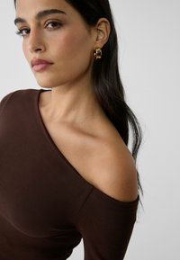 Braunes schulterfreies Oberteil aus einem weichen Stoff. Verfügt über eine elegante Silhouette und dezente Nahtdetails. Akzentuiert mit goldenen Ohrhängern.