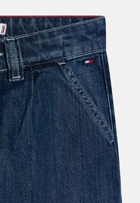 Jeans de mezclilla azul oscuro con una textura suave, que presentan un detalle en rojo en la cinturilla y un pequeño logo de bandera cerca del bolsillo.