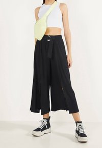 Pantalones de pierna ancha negros con pliegues verticales y aberturas laterales, combinados con una camiseta de tirantes corta blanca y un bolso de bandolera verde claro.