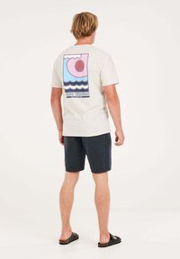 Camiseta de color beige claro con un gráfico colorido de olas y un sol en la parte posterior. Combinada con pantalones cortos oscuros y sandalias negras.