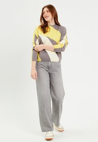 Pull en tricot avec des motifs géométriques jaunes, gris et blancs, col rond et poignets côtelés, associé à un jean large gris et des baskets.