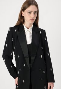 Čierny blazer so saténovými klopami, dvojradové prevedenie, s kovovými ozdobami po celom obvode, kombinovaný s bielou košeľou pod ním.