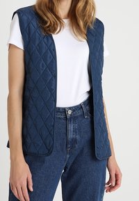 Granatowa, pikowana kamizelka w diamentowy wzór, z otwartym przodem, noszona na białej koszulce, zestawiona z dżinsami o wysokim stanie w kolorze niebieskim. Miękka tekstura.