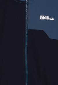 Veste bleu marine avec un panneau contrastant plus clair, fermeture à glissière intégrale et logo Jack Wolfskin en blanc. Texture lisse avec des coutures soignées.