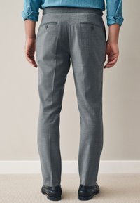 Pantalon gris sur mesure avec une texture lisse, deux poches arrière avec boutons, et une coupe droite. Porté avec des chaussures noires.