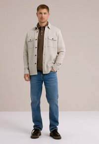 Svetlo siva jakna s gumbom in dvema žepoma na prsih, nosena čez rjavo polo majico, v kombinaciji s srednje modrimi denim hlačami in temnimi čevlji.