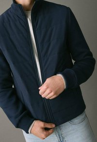 Osoba nosi tamnoplavu kviltanu jaknu preko svijetlosive majice, zatvara jaknu, a vidljive su svijetloplave traperice.