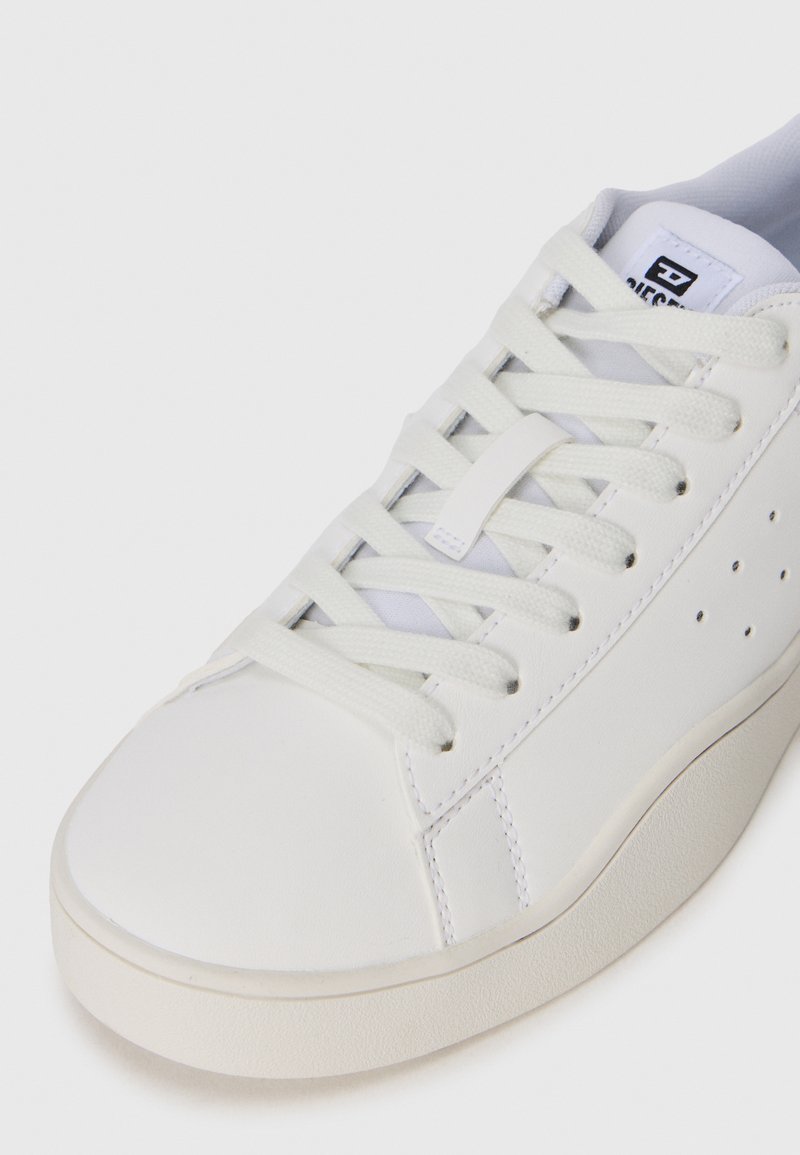 Baskets blanches à tige basse en cuir lisse. Elles ont des lacets plats, un design perforé sur le côté et une épaisse semelle en caoutchouc.