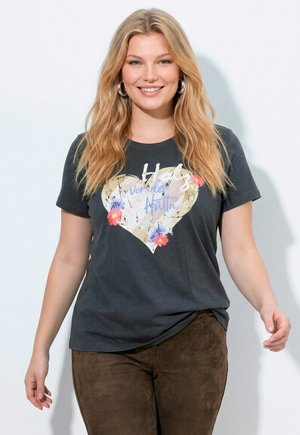 Smilende kvinde med langt blond hår iført en mørkegrå t-shirt med et hjerte- og blomstermotiv og brune bukser, stående foran en ensfarvet baggrund.