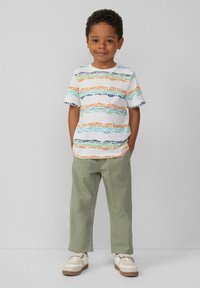 Weißes T-Shirt mit bunten Textmustern in Orange, Blau, Grün und Schwarz, kombiniert mit hellgrünen Hosen und weißen Sneakern mit braunen Sohlen.