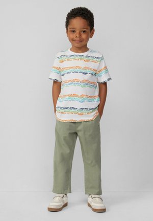 Weißes T-Shirt mit bunten Textmustern in Orange, Blau, Grün und Schwarz, kombiniert mit hellgrünen Hosen und weißen Sneakern mit braunen Sohlen.