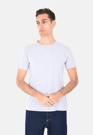 Uomo con maglietta grigio chiaro a maniche corte e jeans blu scuro, in piedi con le mani giunte, guarda leggermente di lato su sfondo bianco.