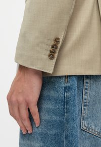 Μπεζ σακάκι με μανίκι που έχει τέσσερα καφέ κουμπιά και λεπτομέρειες ραφής, συνδυασμένο με τζιν ύφασμα. Λείος υφασμάτινος συνδυασμός, με κομμένη εφαρμογή.