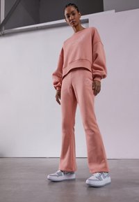 Rosa Sweatshirt und ausgestellte Sweatpants im Set, aus strukturiertem Stoff mit "Be Kind" Stickerei. Kombiniert mit grauen Sneakers.