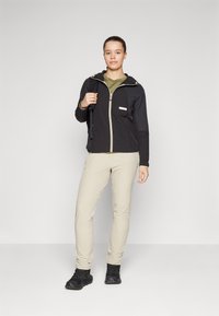 Veste zippée noire avec capuche, accents beiges et poche ; chemise verte olive en dessous, pantalon beige clair et chaussures de randonnée noires.