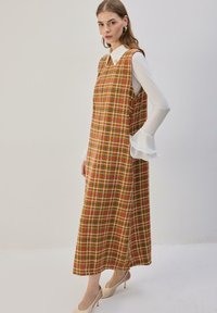 Robe marron à carreaux avec des motifs à grille blancs, roses et bleus. Design sans manches sur une chemise blanche avec des poignets à volants. Coupe jusqu'au sol.