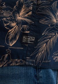 Navy shirt met een beige bloemenpatroon, voorzien van een geweven label. De stof lijkt glad; het ontwerp bevat grote bladeren en een subtiel landschap.