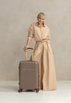 Beige hard-shell suitcase with gold "SUITSUIT" logo, retractable handle, and four wheels; background is neutral with soft texture.
