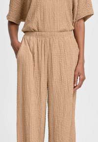 Beige Texturierter Jumpsuit mit kurzen Ärmeln, weitem Bein, elastischem Bund und Seitentaschen. Der Stoff hat eine zerknitterte Optik.