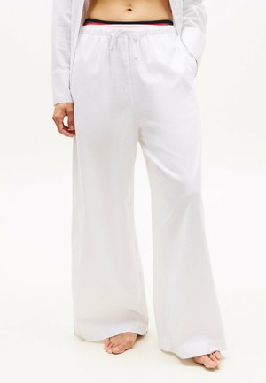 Persona che indossa pantaloni bianchi larghe con coulisse e una fascia in vita con strisce rosse, bianche e blu, in piedi scalza su uno sfondo chiaro.