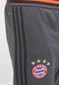 Grijze sportbroek met zwarte zijstrepen, elastische tailleband en het FC Bayern München-embleem in rood en blauw, met gouden sterren erboven.