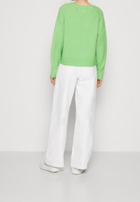 Personne vue de dos portant un pull vert clair, un pantalon blanc à jambes larges et des baskets blanches, sur un fond uni.