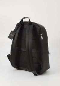 Sac à dos noir avec des panneaux en cuir lisse et en maille texturée. Comprend des sangles rembourrées, une poignée supérieure et une fermeture à glissière. Inclut une étiquette.