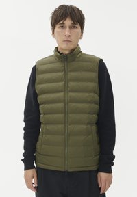 Moški nosi zeleno prešito brezrokavnik preko črne majice z dolgimi rokavi, stoji ob preprosti beli ozadju.