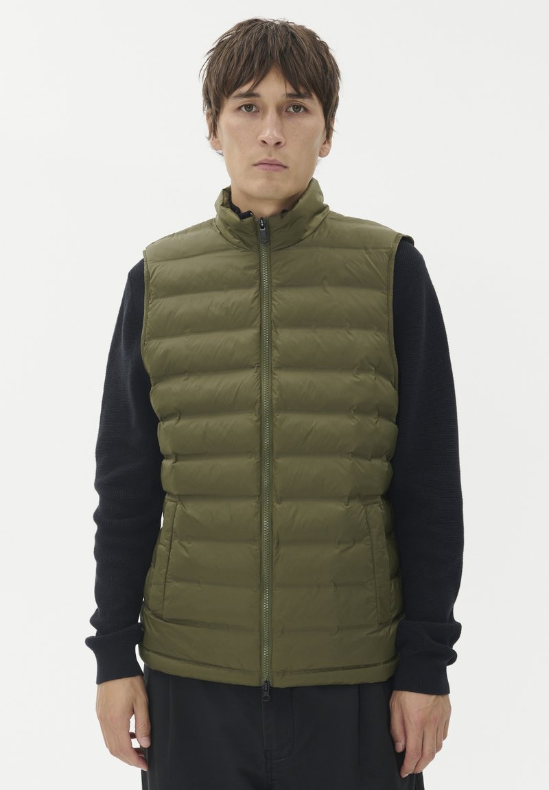 Moški nosi zeleno prešito brezrokavnik preko črne majice z dolgimi rokavi, stoji ob preprosti beli ozadju.