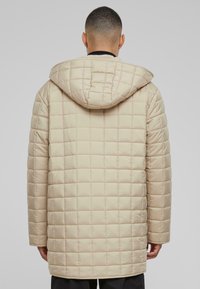 Beige kviltet pufferjakke med hætte, der har et ternet mønster og en glat tekstur, der når til midten af låret.