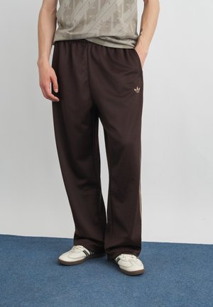 Mand iført mørkebrune Adidas træningsbukser, beige Adidas t-shirt og hvide sneakers, stående på blåt tæppe mod ensfarvet baggrund.