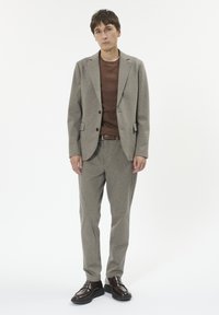 Jachetă de costum gri deschis cu o textură subtilă și pantaloni asortați, pulover tricotat maro dedesubt și pantofi marron slip-on.