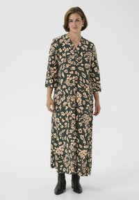 Langes grünes Kleid mit floralem Muster in Beige, mit V-Ausschnitt, 3/4-Ärmeln und lockerer Passform, kombiniert mit schwarzen Knöchelstiefeln.
