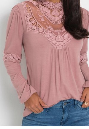 Donna che indossa una maglia a maniche lunghe rosa polvere con dettagli in pizzo sul petto e sulle maniche abbinata a jeans blu, mani nelle tasche.
